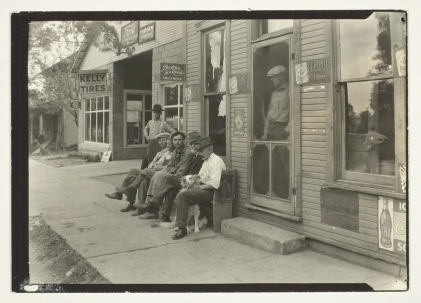 Men With Dog Sitting On Bench In Front Of General Store